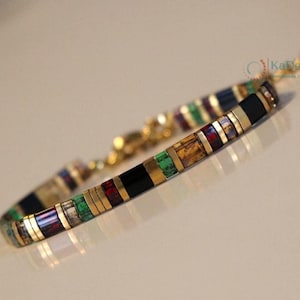 Puede incluir: Una pulsera colorida con detalles dorados. La pulsera presenta segmentos rectangulares en negro, dorado, verde, rojo y azul. El cierre y la cadena de conexión son dorados. La pulsera está diseñada para una muñeca.
