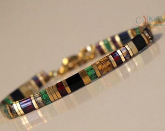 May include: A colorful bracelet with gold accents. The bracelet features rectangular segments in black, gold, green, red, and blue. The clasp and connecting chain are gold. The bracelet is designed for a wrist.