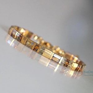 Op de afbeelding: Een goudkleurige armband met rechthoekige segmenten. De armband heeft afwisselende secties van goud en rechthoekige, veelkleurige kralen. De armband wordt gepresenteerd op een reflecterend oppervlak.