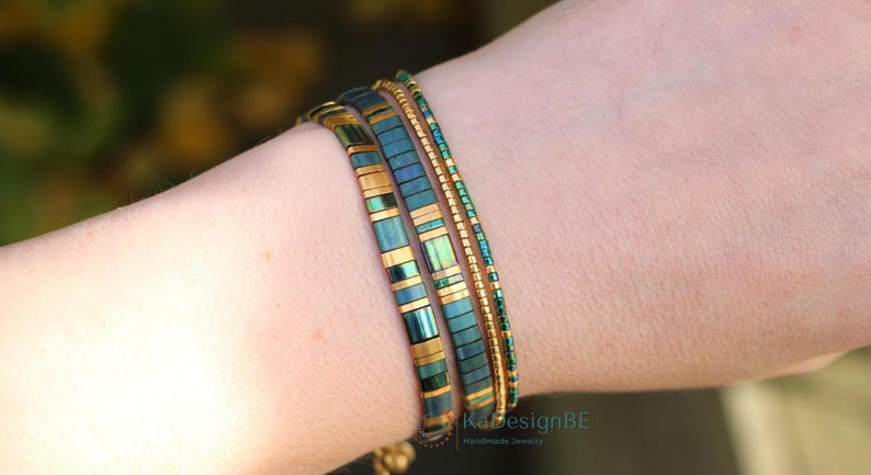 May include: Three bracelets on a wrist. The bracelets are teal and gold. The first bracelet is made of rectangular teal and gold beads. The second bracelet is made of small teal beads. The third bracelet is made of small gold beads.