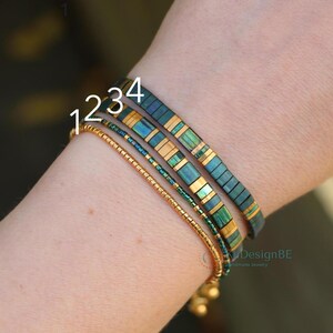 May include: Four bracelets are displayed on a wrist. The bracelets include a gold beaded bracelet, a teal and gold bracelet, a green and gold bracelet, and a dark teal and gold bracelet. The bracelets are handmade jewelry.