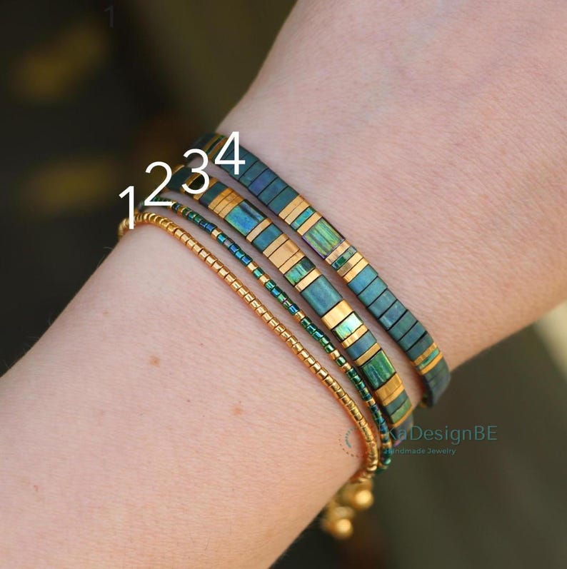 May include: Four bracelets are displayed on a wrist. The bracelets include a gold beaded bracelet, a teal and gold bracelet, a green and gold bracelet, and a dark teal and gold bracelet. The bracelets are handmade jewelry.