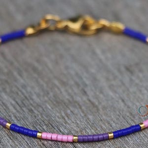 Pode incluir: Um bracelete delicado feito com pequenas contas rosa, roxas e azuis, intercaladas com detalhes dourados. O bracelete tem um fecho dourado.
