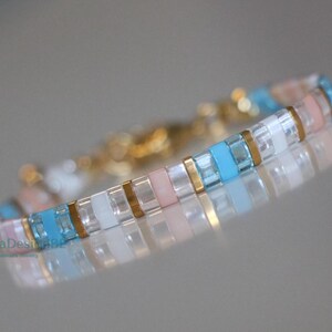 May include: A delicate bracelet featuring a repeating pattern of small, rectangular beads in pastel pink, light blue, white, and gold. The bracelet is made of small, rectangular beads and has a gold clasp.