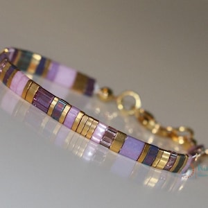 Op de afbeelding: Een paars-gouden kralenarmband met een gouden kettingsluiting. De armband is gemaakt van kleine, vierkante kralen in tinten paars, roze en goud.