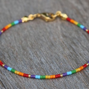 May include: A delicate bracelet featuring a rainbow of small, colorful beads. The beads are arranged in a linear pattern, transitioning from red to orange, yellow, green, blue, and purple. Gold-colored accents and clasp complete the design.