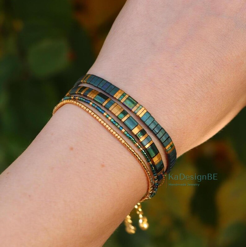 May include: A set of four bracelets in shades of gold, teal, and dark blue. The bracelets are worn on a wrist. The top two bracelets have a rectangular design with gold accents. The bottom two bracelets are made of small beads. The text "KaDesignBE" is visible.