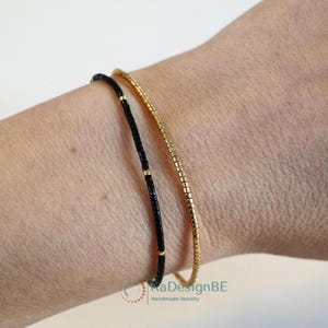 Op de afbeelding: Twee delicate armbanden om een pols. Eén is zwart met gouden accenten, de andere is goudkleurig. De armbanden zijn dun en minimalistisch, geschikt voor dagelijks gebruik. De sieraden zijn handgemaakt.