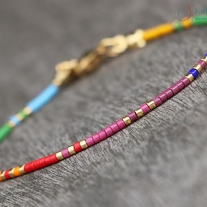 Peut inclure: Un bracelet délicat aux couleurs de l'arc-en-ciel, composé de perles rectangulaires rouges, oranges, jaunes, vertes, bleues et violettes. Il est orné d'accents dorés et d'un fermoir doré. Le bijou est présenté sur une surface grise.
