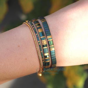 May include: A collection of bracelets in shades of gold and teal. The bracelets are worn on a wrist and include a delicate gold chain, a beaded strand, and two wider bands with rectangular teal and gold accents. The background is blurred.