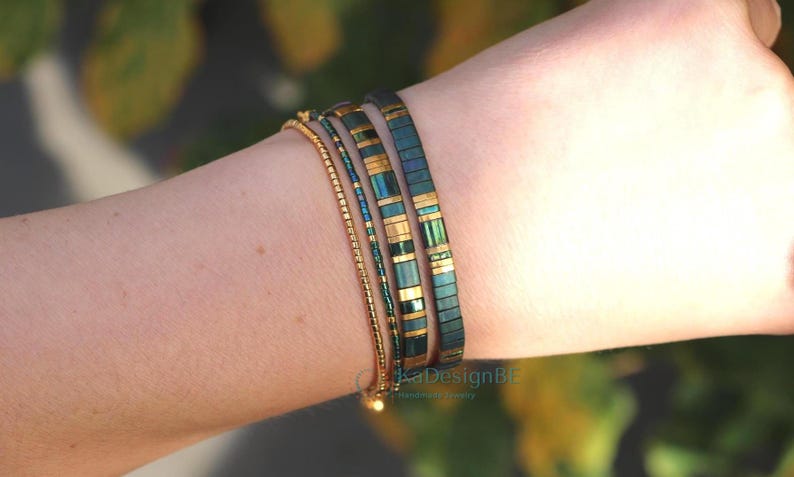 May include: A collection of bracelets in shades of gold and teal. The bracelets are worn on a wrist and include a delicate gold chain, a beaded strand, and two wider bands with rectangular teal and gold accents. The background is blurred.