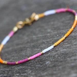 Könnte beinhalten: Ein filigranes Perlenarmband mit farbenfrohem Design. Das Armband zeigt kleine, zylindrische Perlen in Orange, Gelb, Weiß, Rosa und Lila. Goldfarbene Metallakzente und ein Verschluss vervollständigen das Design. Das Armband ist ca. 17 cm lang.