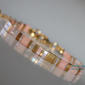Könnte beinhalten: Ein zartes Armband mit rosa, weißen und goldenen Perlen. Das Armband besteht aus kleinen, quadratischen Perlen, die in einem sich wiederholenden Muster aufgereiht sind. Das Armband hat einen goldenen Verschluss.