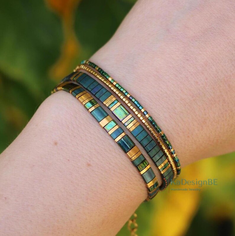 May include: A set of four bracelets in shades of teal, gold, and green. The bracelets are worn on a wrist and feature a mix of rectangular and beaded designs. The bracelets have a delicate and elegant appearance.