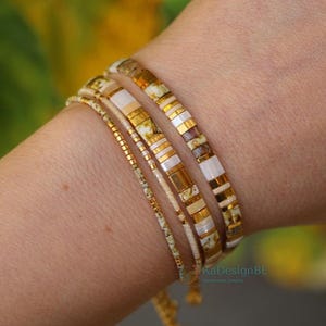 Op de afbeelding: Vier armbanden worden getoond op een pols. De armbanden hebben gouden, witte en patroon kralen. Eén armband is een delicate gouden ketting. De armbanden zijn handgemaakte sieraden.