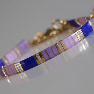 May include: A delicate bracelet with a repeating pattern of flat, rectangular beads in shades of purple, lavender, and blue. Gold-toned metal accents and a gold chain clasp complete the design. The bracelet is a piece of jewellery.