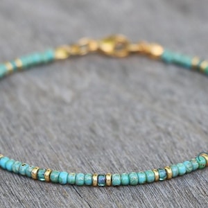 May include: A delicate turquoise and gold beaded bracelet with a clasp closure. The bracelet is made with small, round beads and features a few larger, square beads.