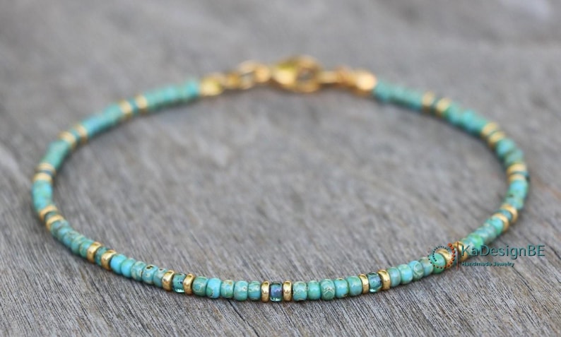 May include: A delicate turquoise and gold beaded bracelet with a clasp closure. The bracelet is made with small, round beads and features a few larger, square beads.