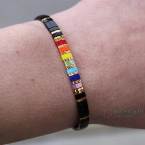 May include: A black and gold bracelet with rainbow colored beads. The bracelet is made of small square beads with gold accents. The beads are arranged in a rainbow pattern, starting with red and ending with purple. The bracelet is worn on a person's wrist.