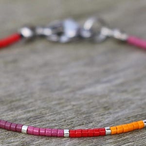 May include: A rainbow beaded bracelet with silver accents. The bracelet is made with small, square beads in a variety of colors, including red, orange, yellow, green, blue, and purple.