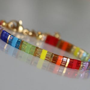 以下が含まれることがあります： ゴールドのアクセントが付いた虹色のビーズブレスレット。ブレスレットは、紫、青、ターコイズ、緑、黄、オレンジ、赤の色合いの小さな正方形のビーズで作られています。