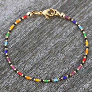 Rainbow anklet in top view on a wooden surface/background. The bracelet has its center down on the picture, the closure (stainless steel lobster clasp) is on top of the picture. The anklet is handmade with the pride colors in following pattern: 2 red, 2 orange, 2 yellow, 2 green, 2 turquoise, 2 cobalt blue, 2 dark purple, 2 light purple beads. In between each different color, there is a pattern of gold-black-gold beads. This tiny bracelet is subtle pride, stylish, dainty, minimalist.