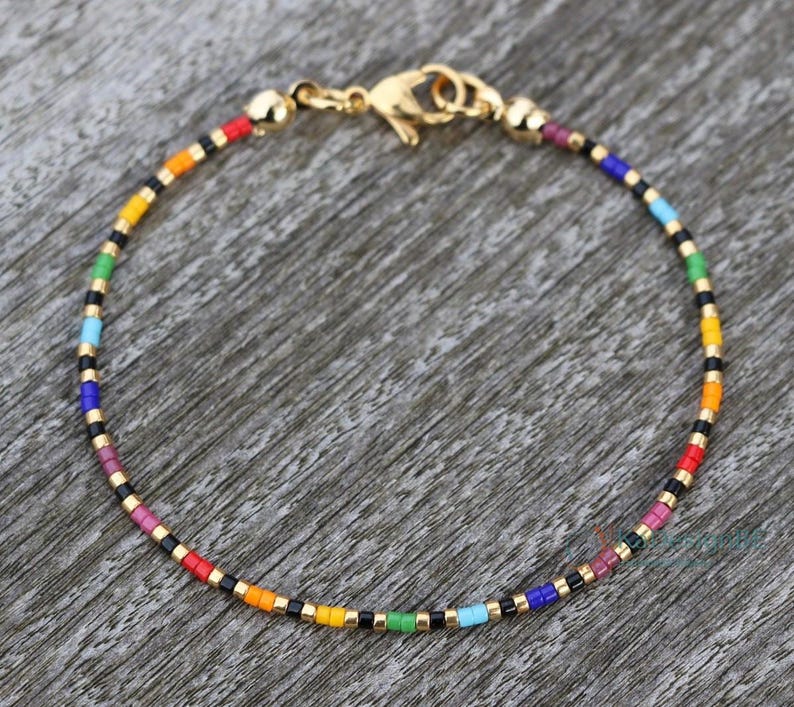 Rainbow anklet in top view on a wooden surface/background. The bracelet has its center down on the picture, the closure (stainless steel lobster clasp) is on top of the picture. The anklet is handmade with the pride colors in following pattern: 2 red, 2 orange, 2 yellow, 2 green, 2 turquoise, 2 cobalt blue, 2 dark purple, 2 light purple beads. In between each different color, there is a pattern of gold-black-gold beads. This tiny bracelet is subtle pride, stylish, dainty, minimalist.