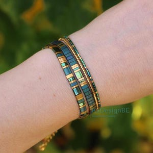 May include: Three beaded bracelets in shades of teal and gold are worn on a wrist. The bracelets have a rectangular and cylindrical bead design. The background is a blurred green and yellow.