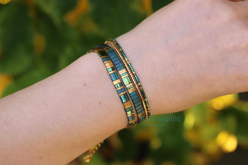 May include: Three beaded bracelets in shades of teal and gold are worn on a wrist. The bracelets have a rectangular and cylindrical bead design. The background is a blurred green and yellow.