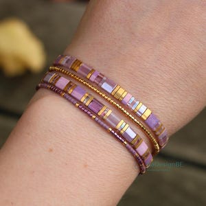 Op de afbeelding: Close-up van drie armbanden gestapeld op een pols. De armbanden hebben een mix van paarse, gouden en roze kralen. Eén armband is gemaakt van kleine paarse kralen, terwijl de andere rechthoekige en goudkleurige accenten hebben.