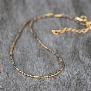 Op de afbeelding: Een delicate ketting met een mix van kleine, gespikkelde kralen en goudkleurige metalen segmenten. De ketting heeft een goudkleurige ketting en sluiting en wordt gepresenteerd op een getextureerd, grijs oppervlak.