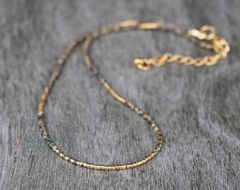 May include: A delicate necklace featuring a mix of small, speckled beads and gold-colored metal segments. The necklace has a gold-toned chain and clasp, and is displayed on a textured, gray surface.