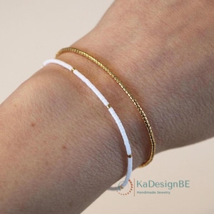 Pode incluir: Duas pulseiras delicadas em um pulso. Uma é uma fina corrente dourada, e a outra é branca com detalhes dourados. As pulseiras são simples e elegantes, adequadas para o uso diário. A imagem é bem iluminada, mostrando os detalhes das joias.