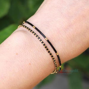 May include: Two delicate bracelets, one above the other, are visible on a wrist. The top bracelet is black, and the bottom bracelet is gold. Both bracelets are made of small beads. The background is a soft green.