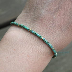K&ouml;nnte beinhalten: Ein t&uuml;rkisfarbenes und goldenes Armband, das an einem Handgelenk getragen wird. Das Armband besteht aus kleinen, runden Perlen mit einem dezenten, gefleckten Muster.