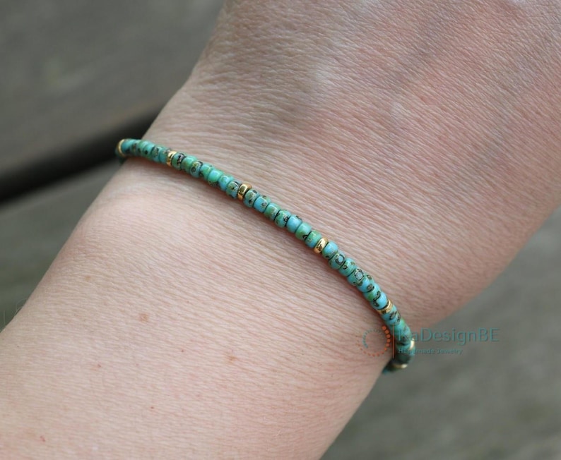 K&ouml;nnte beinhalten: Ein t&uuml;rkisfarbenes und goldenes Armband, das an einem Handgelenk getragen wird. Das Armband besteht aus kleinen, runden Perlen mit einem dezenten, gefleckten Muster.