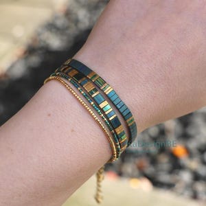May include: A collection of three bracelets. Two bracelets feature a teal and gold rectangular design, while the third is a delicate gold beaded chain. The bracelets are worn together on a wrist.