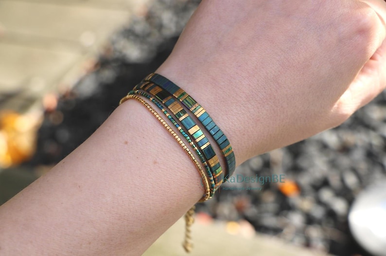 May include: A collection of three bracelets. Two bracelets feature a teal and gold rectangular design, while the third is a delicate gold beaded chain. The bracelets are worn together on a wrist.
