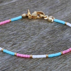 Könnte beinhalten: Ein zartes Armband aus kleinen, bunten Perlen in den Farben Rosa, Weiß und Blau. Das Armband hat einen goldenen Verschluss.