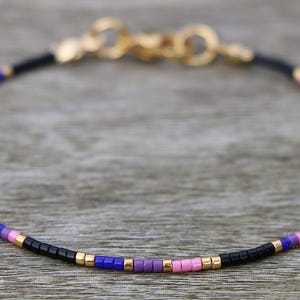 Pode incluir: Um bracelete delicado com contas pretas, cor-de-rosa e roxas. O bracelete tem um fecho dourado.