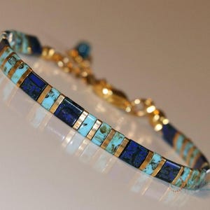 May include: A gold bracelet featuring alternating turquoise and dark blue rectangular stones, separated by gold bars. The bracelet has a gold chain and clasp. The design is elegant and the colours are vibrant.