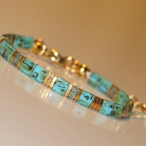 Op de afbeelding: Een turquoise en gouden kralen armband met een sluiting. De armband is gemaakt van kleine, vierkante kralen met een turquoise kleur en gouden accenten.