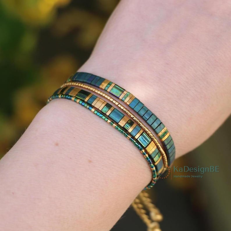 May include: Two beaded bracelets in shades of teal, gold, and green are stacked on a wrist. The bracelets feature rectangular and round beads. The bracelets are handmade jewelry.