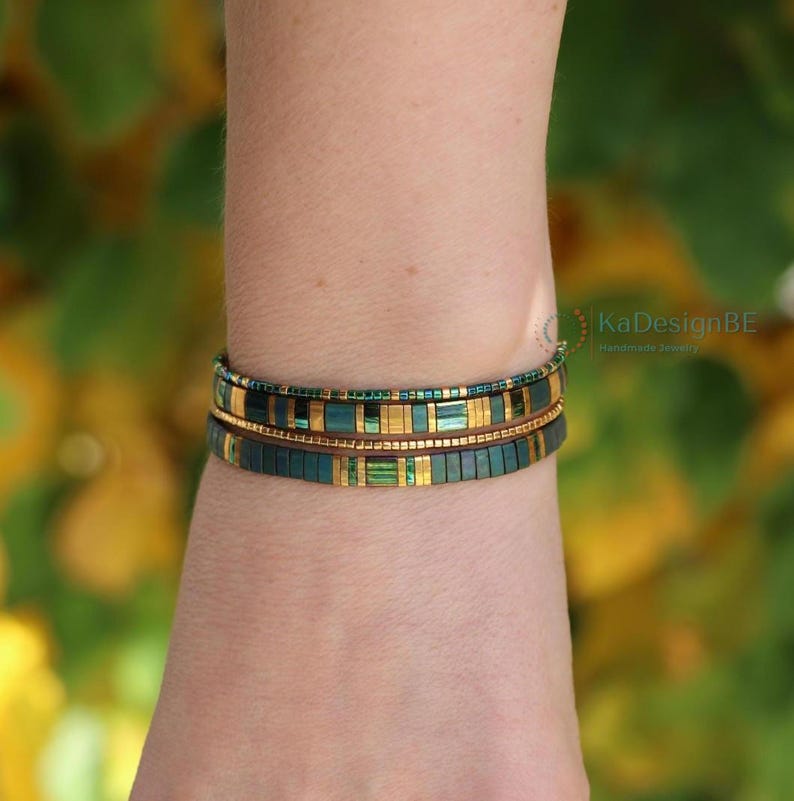 May include: Two beaded bracelets in shades of teal, gold, and green. The bracelets are made of small, rectangular beads and are stacked on top of each other. The bracelets are handmade jewelry, as indicated by the text "KaDesignBE Handmade Jewelry".