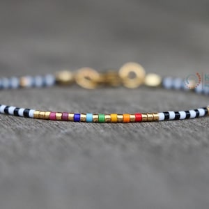 Op de afbeelding: Een delicate kralenarmband met een regenboog aan kleuren en gouden accenten. De armband wisselt af tussen zwart-wit gestreepte kralen en gouden kralen. De sluiting is goudkleurig. De armband ligt op een grijze ondergrond.