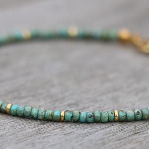 K&ouml;nnte beinhalten: Ein t&uuml;rkisfarbenes und goldenes Perlenarmband mit einem Verschluss. Das Armband besteht aus kleinen, runden Perlen und wirkt zart.