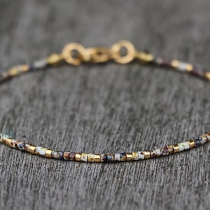 Op de afbeelding: Delicate kralenarmband met een gouden sluiting. De armband heeft kleine, kleurrijke kralen in tinten goud, wit, bruin en marineblauw. De armband is ongeveer 18 cm lang.