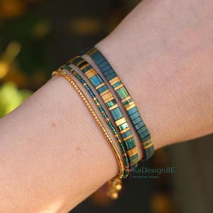 May include: A collection of handmade bracelets in shades of teal, gold, and green. The bracelets include a delicate gold chain, a beaded bracelet, and two flat, rectangular tile bracelets. The bracelets are stacked on a wrist.