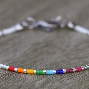 Peut inclure: Un bracelet délicat composé de petites perles blanches avec un arc-en-ciel de perles colorées au centre. Le bracelet a un fermoir argenté et mesure environ 18 cm de long.