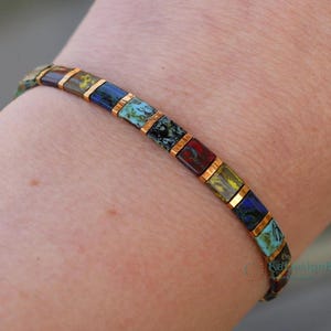 Op de afbeelding: Een kleurrijke armband met rechthoekige kralen in tinten blauw, groen, rood en geel, gescheiden door goudkleurige metalen schakels. De armband wordt om een pols gedragen.